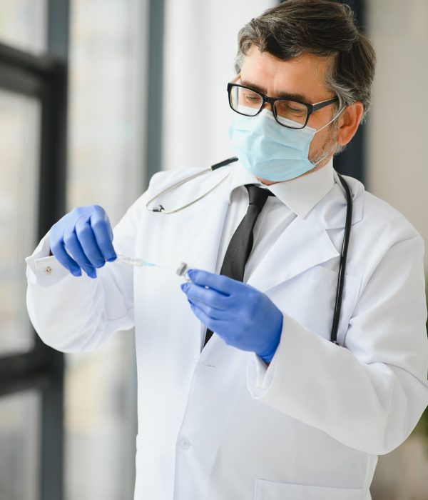 Male doctor wearing uniform, mask, medical gloves holding syringe